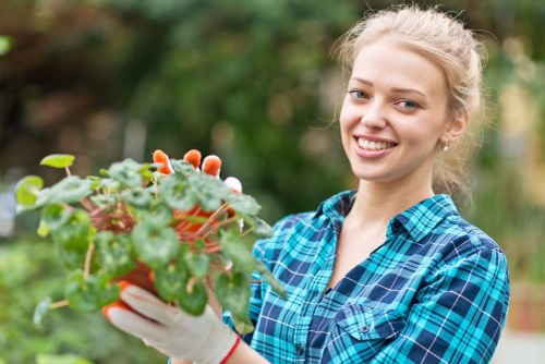 A young woman with blonde hair tied back, wearing a blue and green plaid shirt and white gardening gloves, is smiling while holding a small potted plant with green leaves and some reddish stems. The background features a blurred garden scene with trees and foliage, indicating an outdoor gardening space. The image illustrates a gardening activity, possibly in the front or back garden of a residential property in Plaistow, highlighting natural outdoor environments suitable for professional gardening and landscaping work by Gardening Services Plaistow.