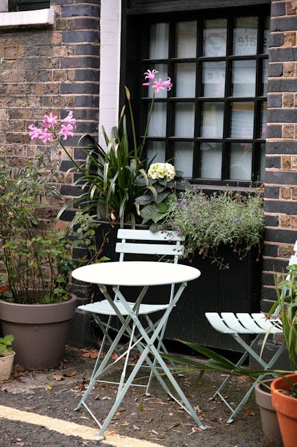 A cozy urban garden corner featuring a white metal bistro table with matching chairs situated on a paved surface. Potted plants, including a large container with a mix of pink flowers and greenery, ad