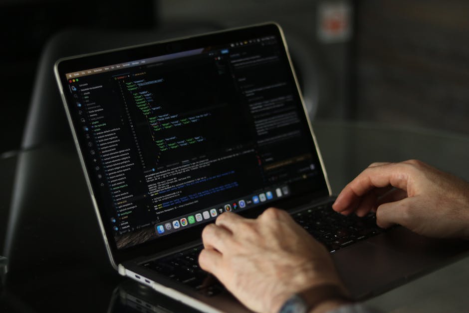 Close-up of a person's hands typing on a MacBook laptop situated on a dark table. The laptop screen displays lines of colorful computer code, indicating programming activity. The background is slightl